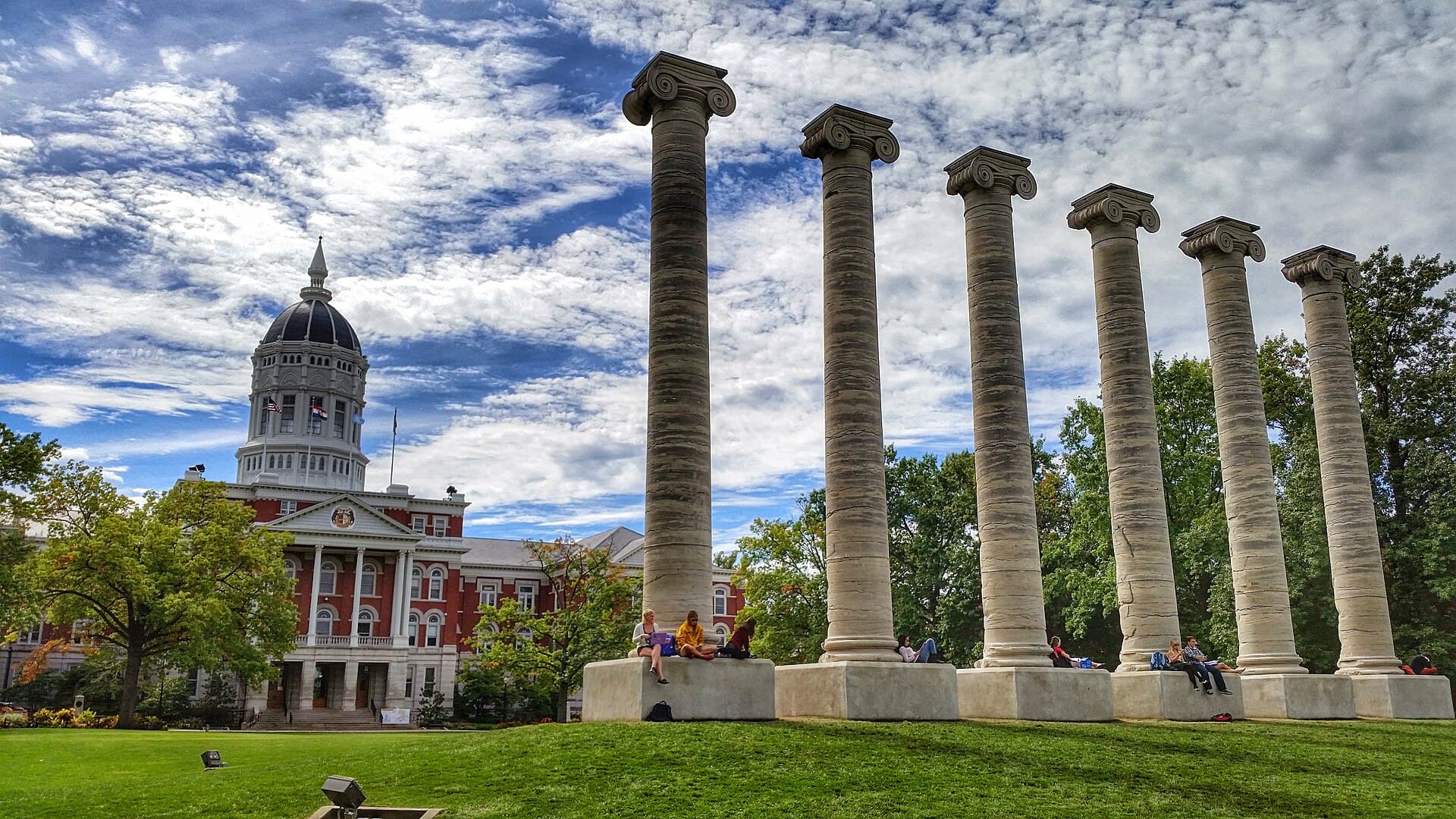 The domed red-brick Jesse Hall beyond the six stone Columns on Francis Quadrangle, with students sitting on the column bases under a cloudy blue sky