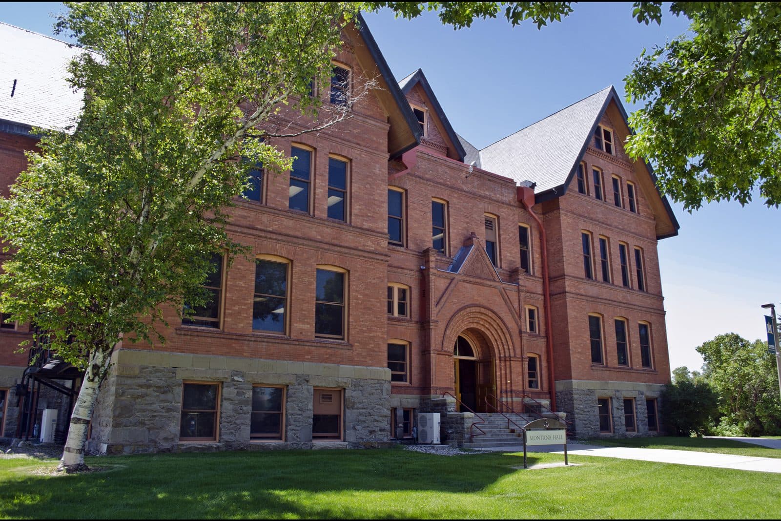 A four-story red-brick hall with a stone base, steep gray slate gables, rows of tall windows and an arched stone entrance, behind a green lawn with a small sign, framed by a birch tree and leafy branches under a clear blue sky