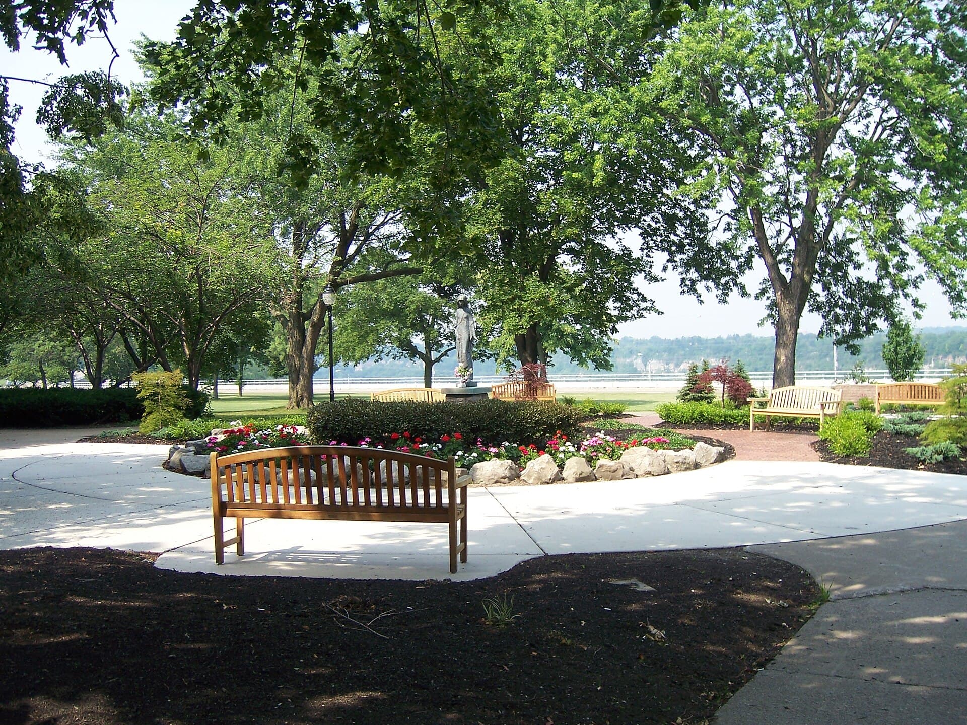 A shady garden at Niagara University with wooden benches around a flower bed and a white statue, tall leafy trees overhead and the wooded far side of the Niagara Gorge in the distance