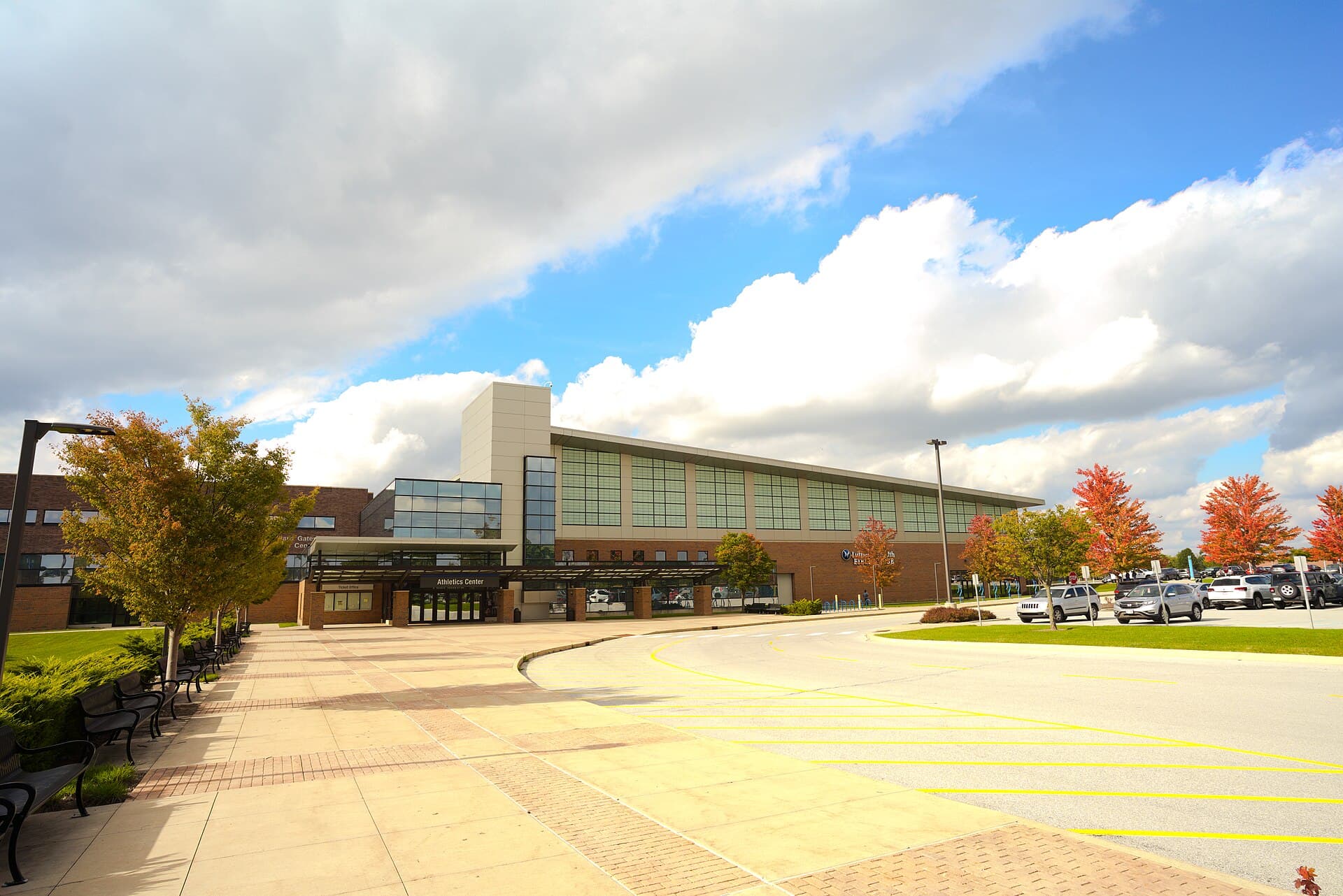 The Hilliard Gates Sports Center and Athletics Center at Purdue Fort Wayne, a long brick-and-glass building with a covered entrance, behind a wide paved plaza and a parking lot lined with red- and orange-leafed trees under a blue sky with big white clouds