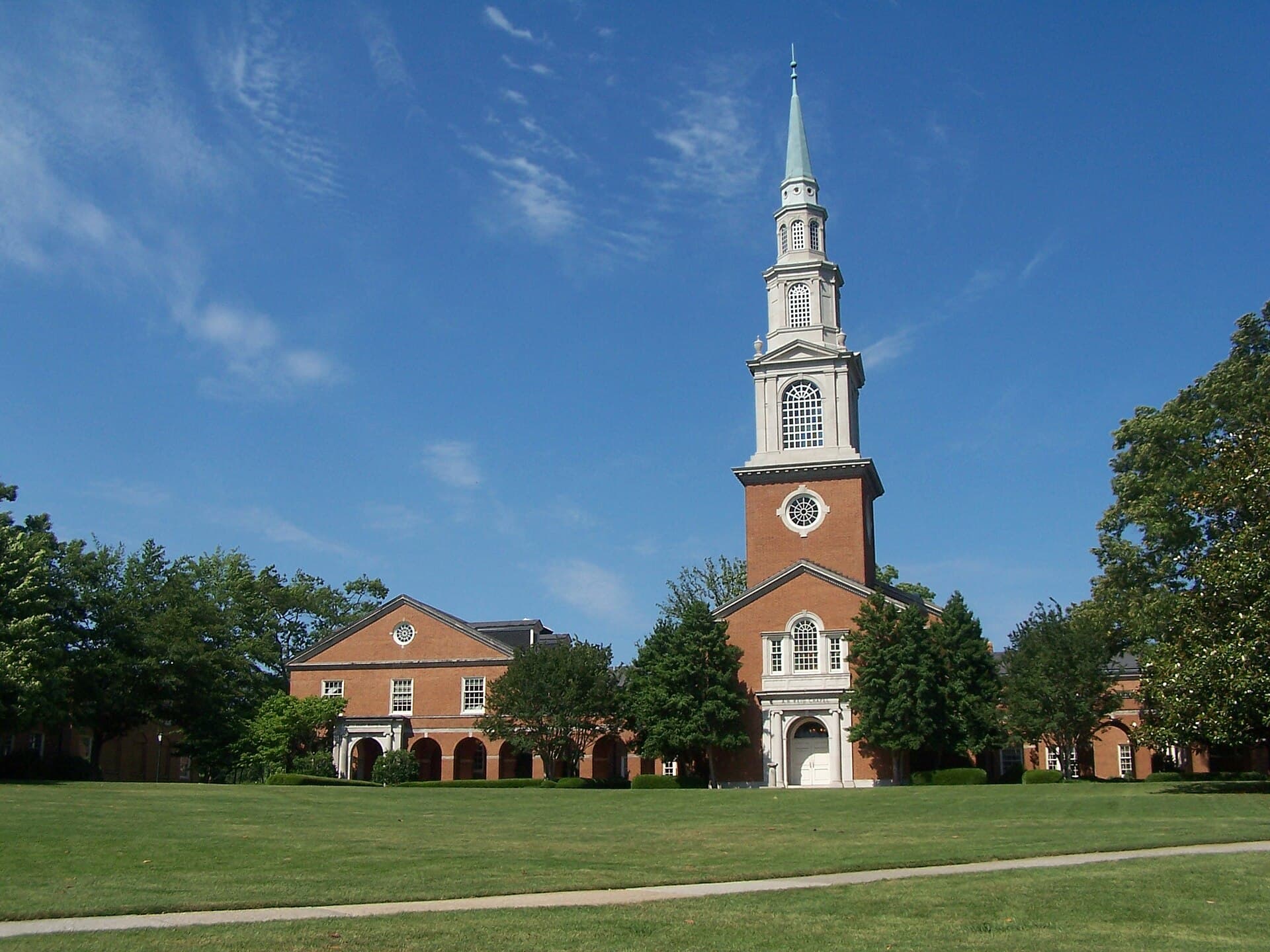 Reid Chapel at Samford University, a red-brick Georgian-style chapel with white columns and a tall white steeple, beyond a green lawn and trees under a blue sky