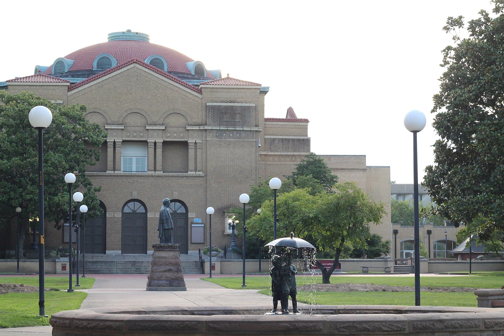 A tan-brick auditorium with a red tile dome and arched doorways behind a bronze statue on a stone pedestal, with a small fountain sculpture of two children under an umbrella in the foreground, globe lampposts and leafy trees under a pale sky