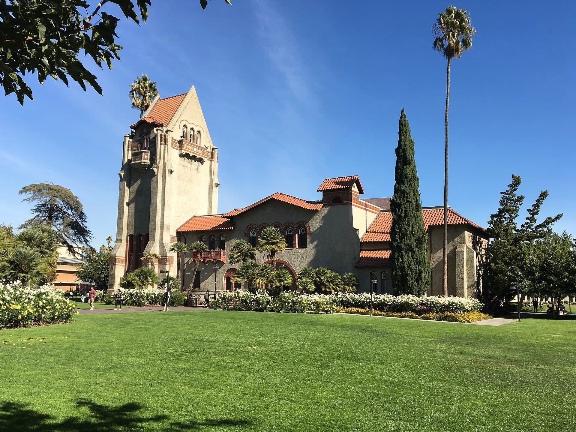 Tower Hall, a tan Mission Revival building with a tall bell tower and red tile roofs, behind a wide green lawn and palm trees