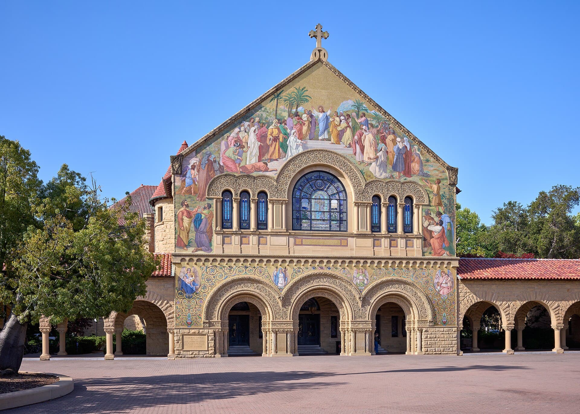 The mosaic-covered sandstone facade of Stanford Memorial Church, with arched entrances, seen from the Main Quad under a clear blue sky