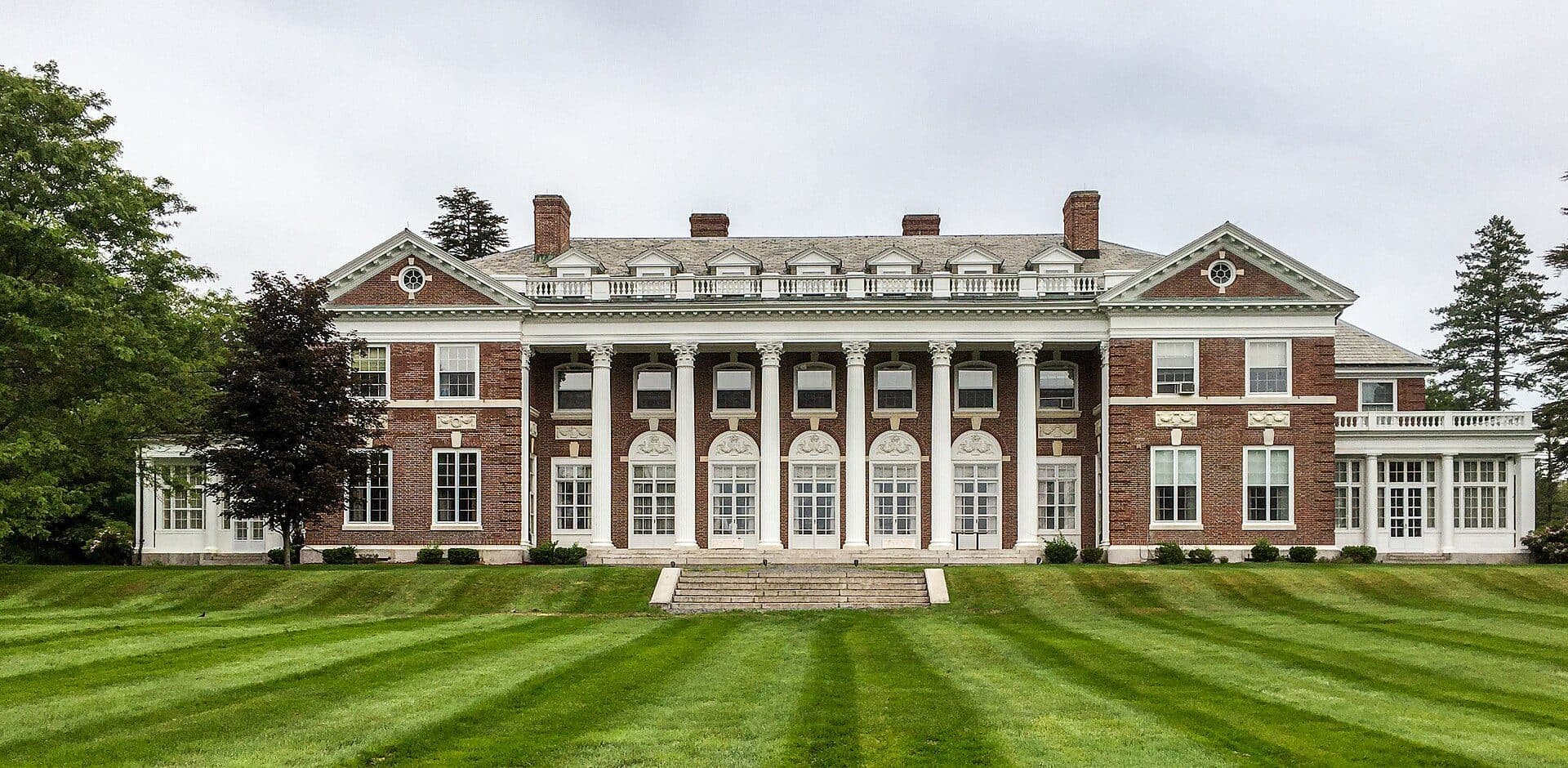 The Georgian-style brick mansion of the former Ames estate at Stonehill College, with white columns and a long row of windows, above a wide striped green lawn and trees