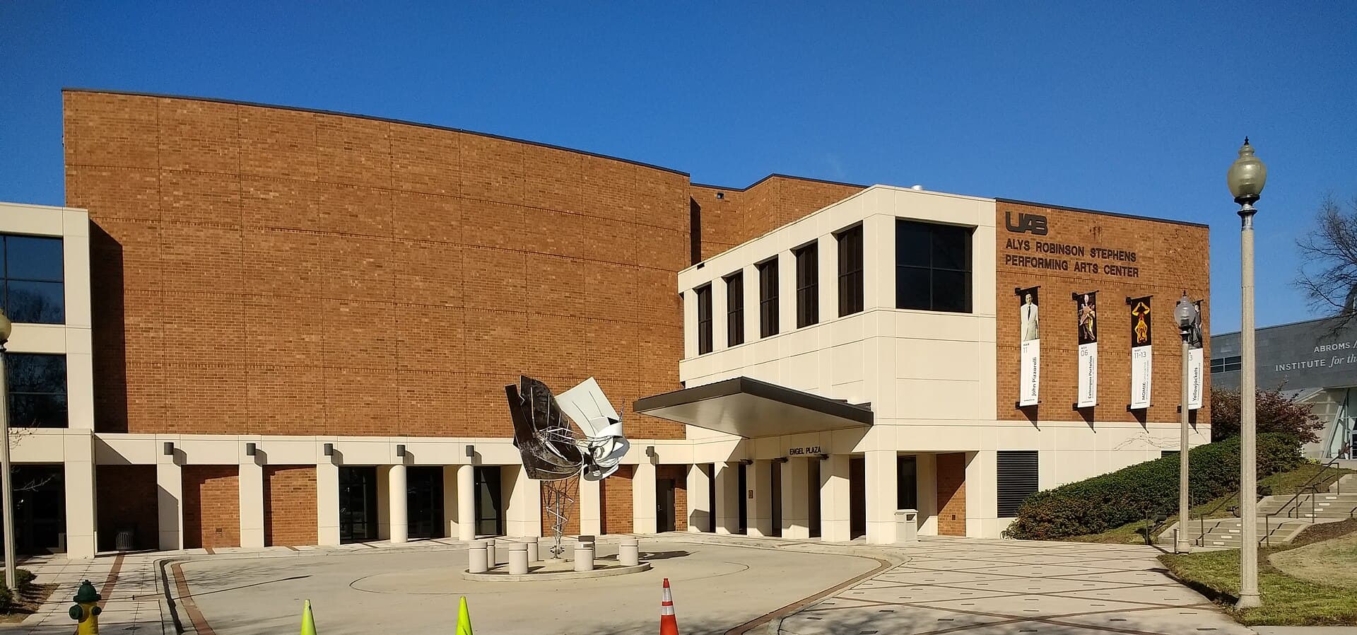 The Alys Robinson Stephens Performing Arts Center at UAB, a long brick building with a white entrance pavilion, a plaza with a modern sculpture and a lamppost under a blue sky
