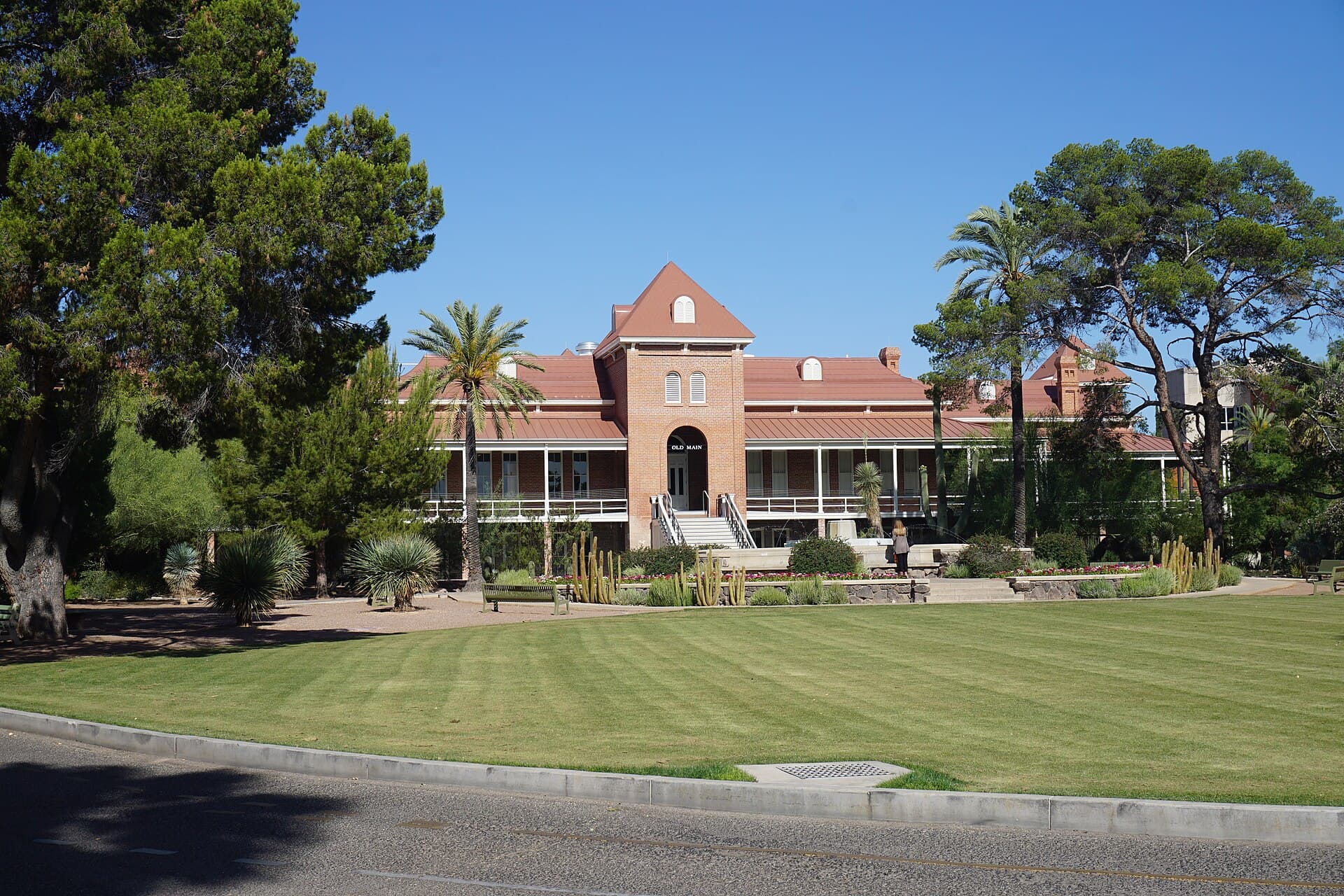 The University of Arizona's red-brick Old Main, with a central gabled tower, wraparound white-railed porches and a red roof, behind a green lawn framed by pine and palm trees under a clear blue sky
