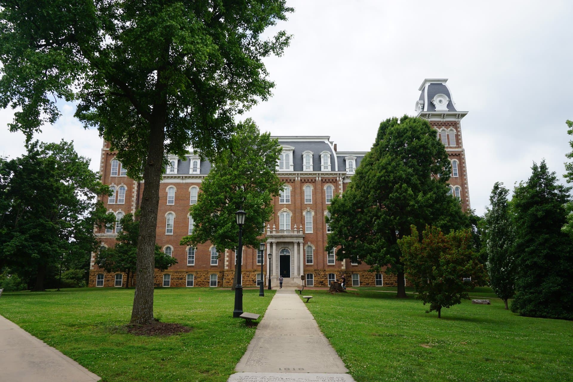 Old Main, a red-brick Victorian building with two mansard-roofed towers at the University of Arkansas, seen down a paved walk between green lawns and tall shade trees under an overcast sky