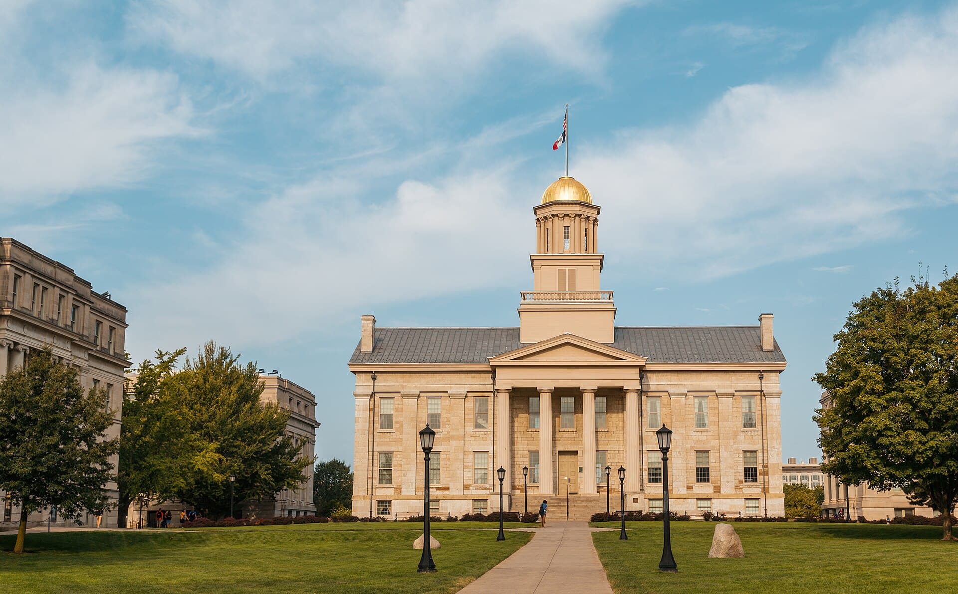 The Greek Revival Old Capitol with its gold dome and columned portico at the end of a lawn and walkway on the Pentacrest, under a blue sky with white clouds