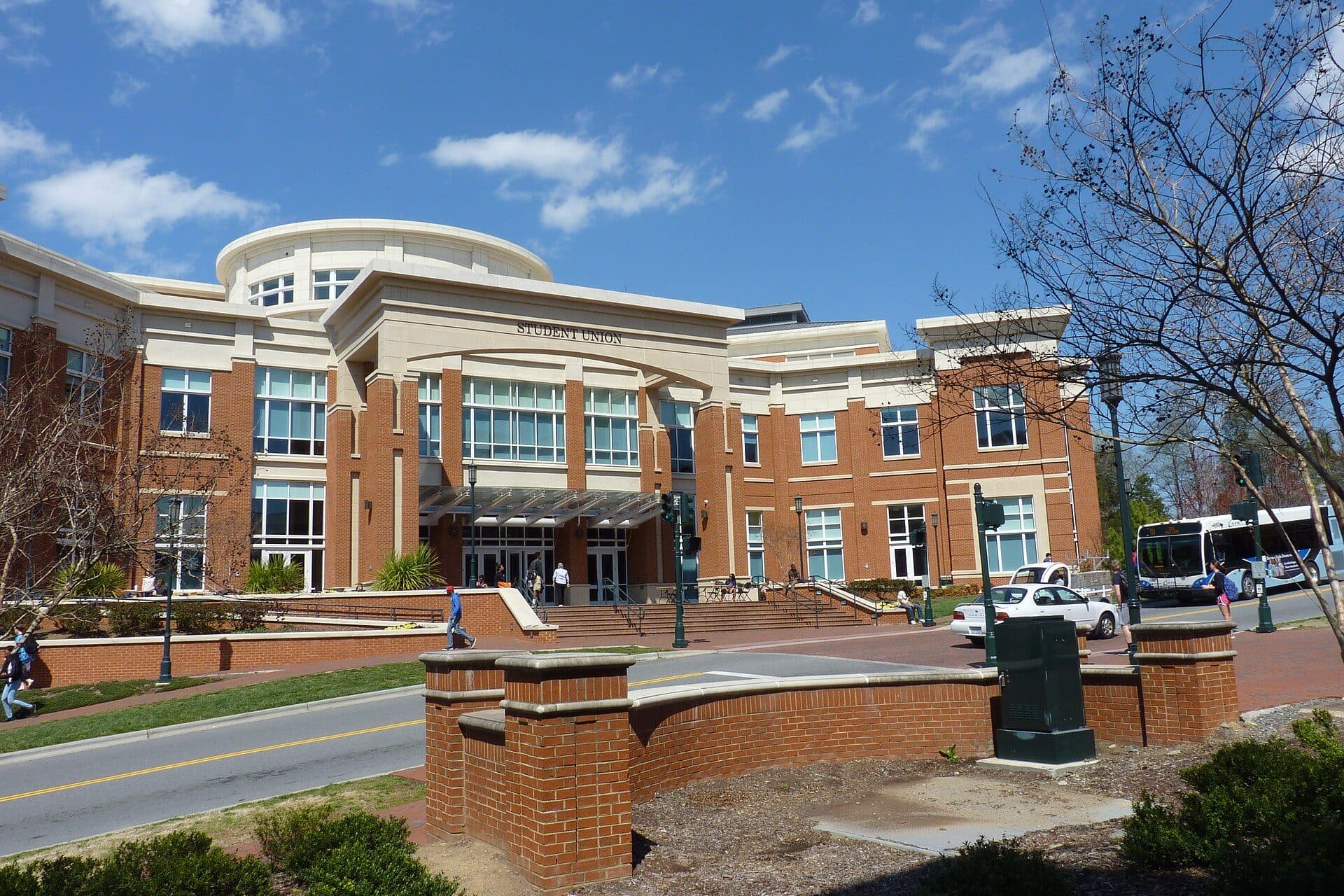 A curved brick-and-glass building on the UNC Charlotte campus with a campus bus out front