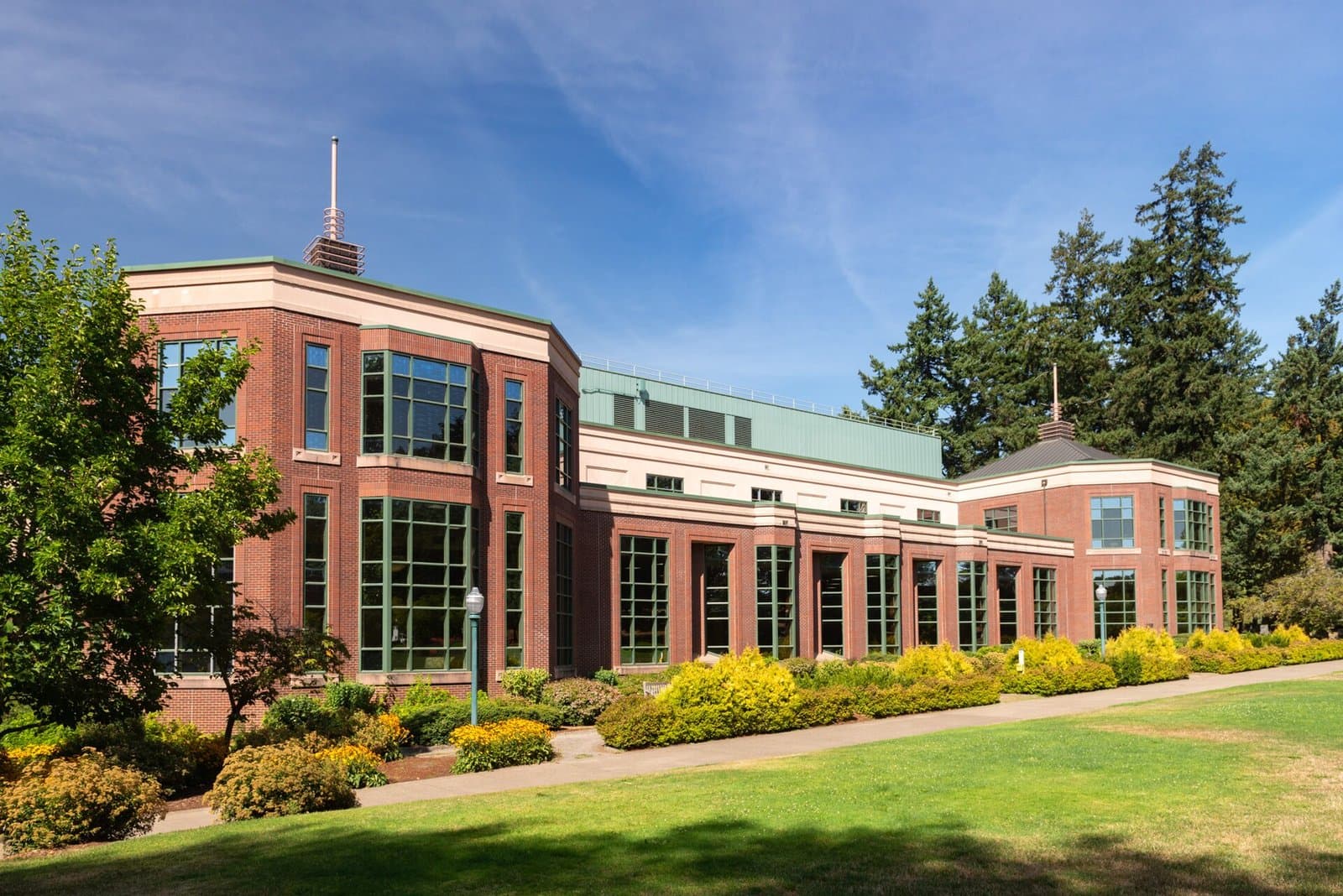 A red-brick wing of the Knight Library with tall green-framed windows above a sunny lawn and flowering shrubs, with tall evergreens behind, at the University of Oregon
