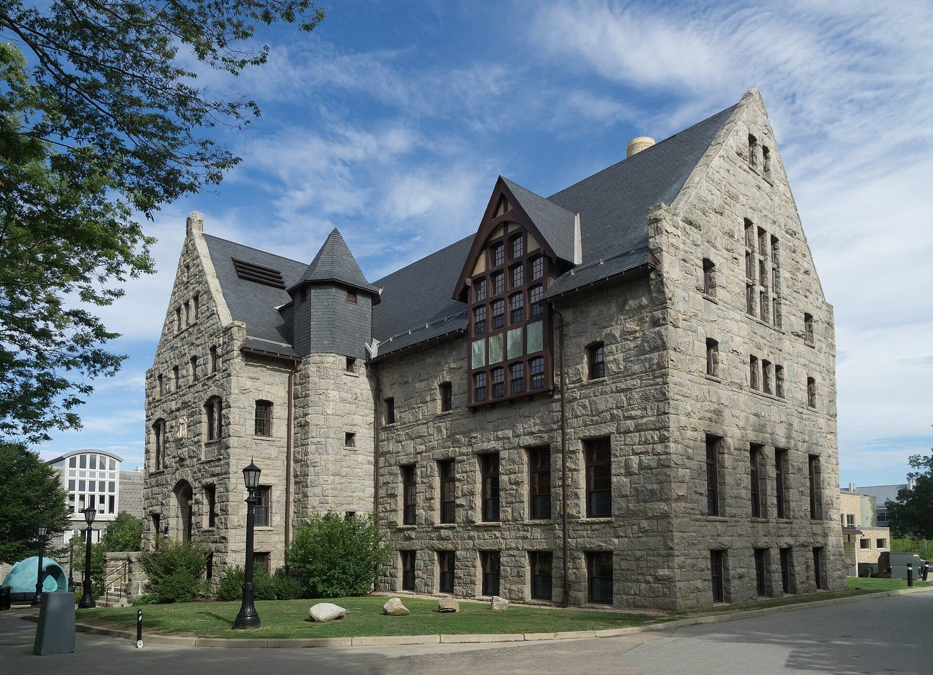 Lippitt Hall at the University of Rhode Island, a gray granite building with steep slate gables, a squat tower and arched windows, under a blue sky with puffy white clouds and a leafy tree at left