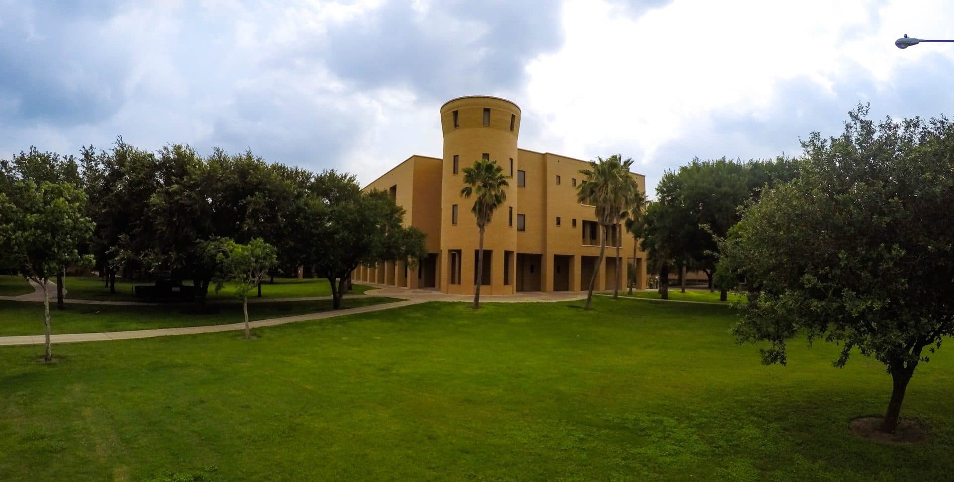 The tan-brick University Library at UTRGV in Edinburg, with a round tower and palm trees, behind a wide green lawn and live oaks under a cloudy sky
