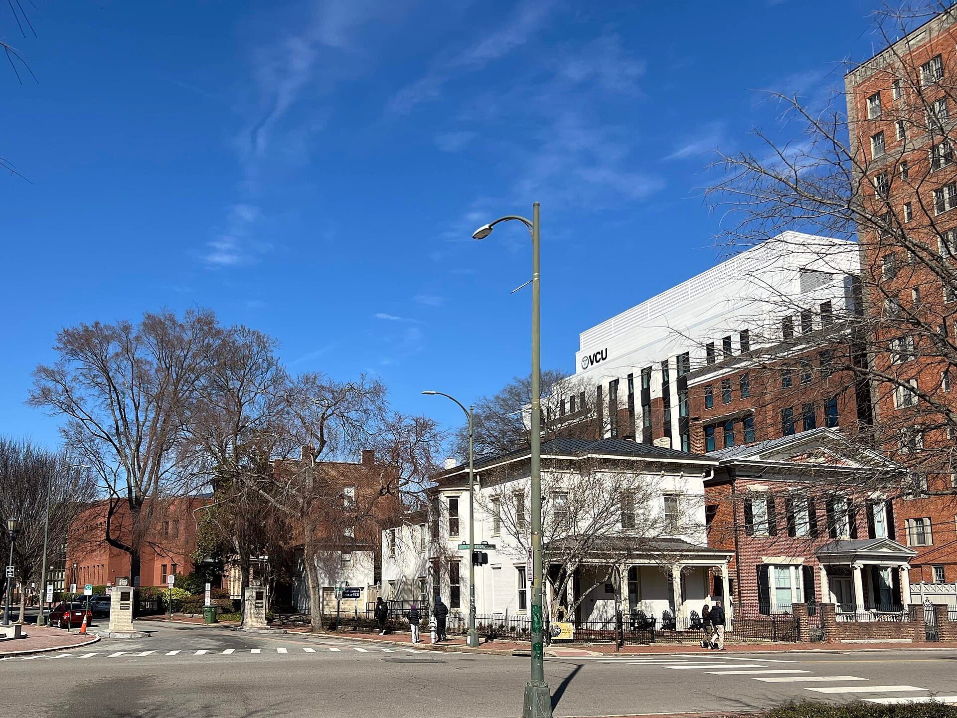 A street corner on VCU's Monroe Park campus with a white historic house, a white building marked VCU and brick buildings behind bare trees under a blue sky