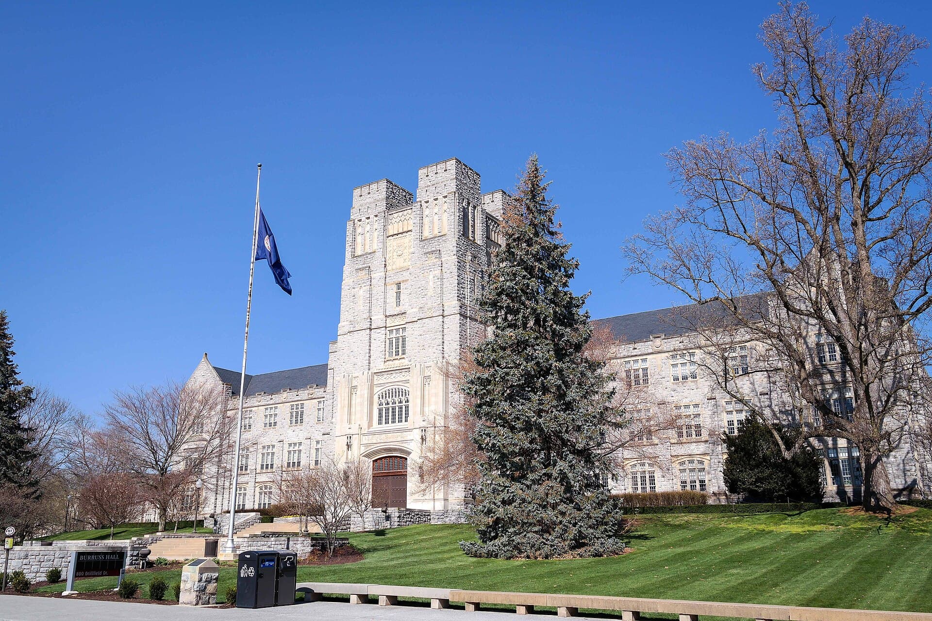 Burruss Hall at Virginia Tech, a Gothic-style limestone building with a central tower, behind a lawn and an evergreen tree under a blue sky