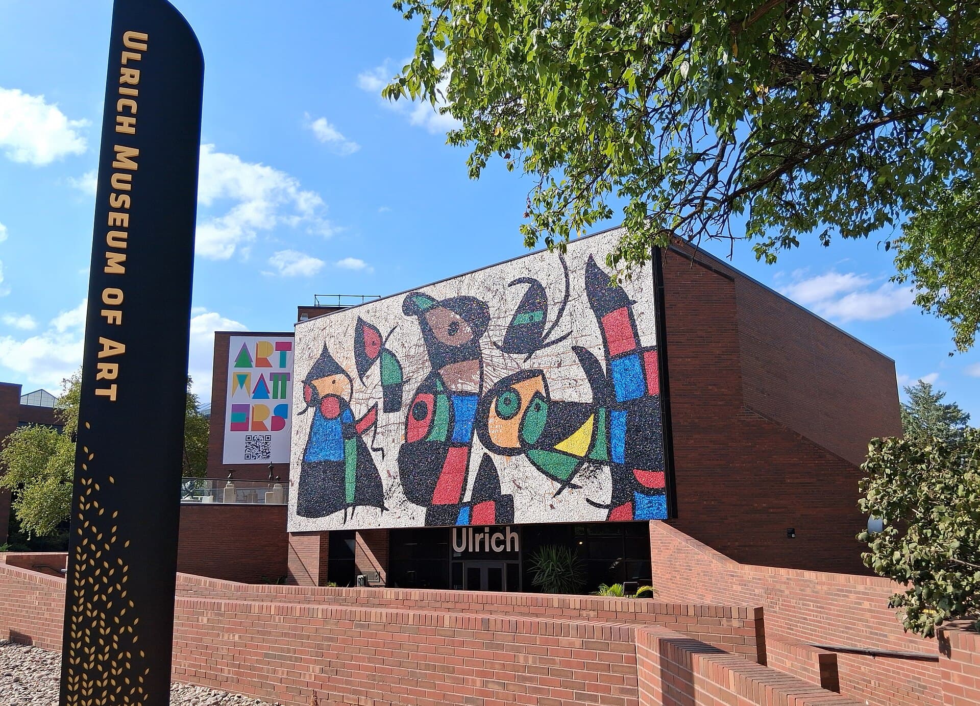 A tall black sign reading ULRICH MUSEUM OF ART in front of a red-brick building whose wall is covered by a large, colorful mosaic mural of abstract bird-like figures, with low brick walls, a tree and a clear blue sky